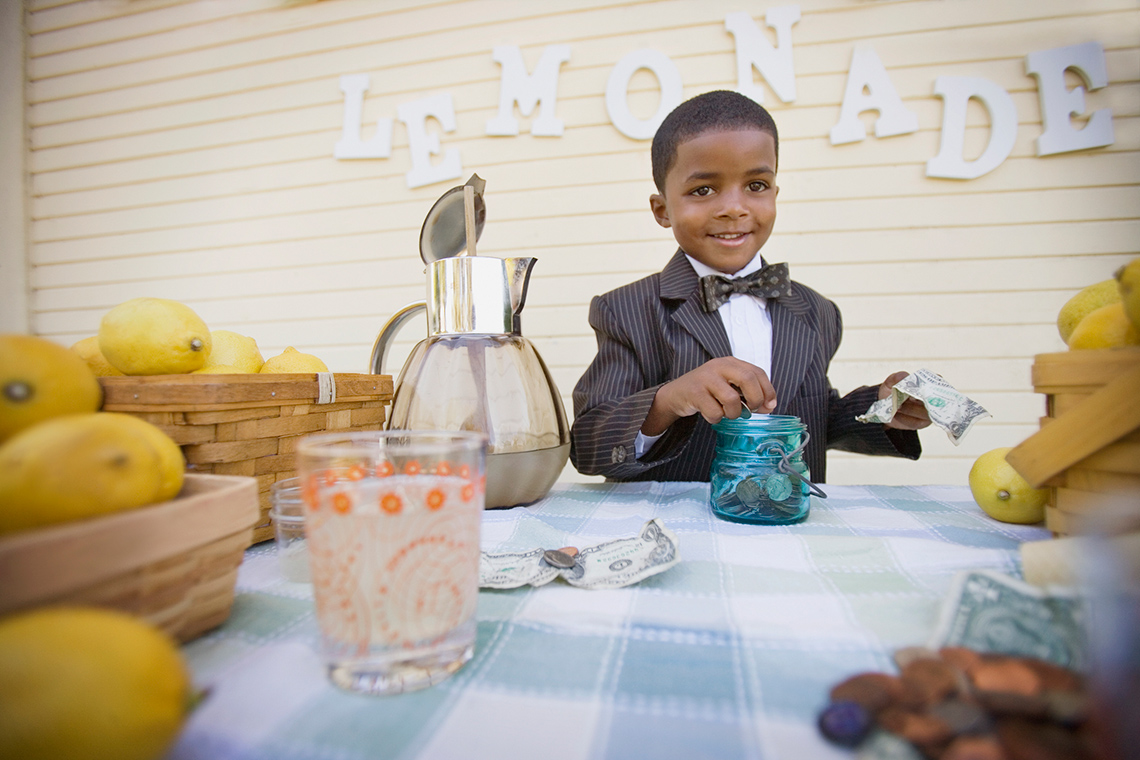 A small child wearing a suit adds money to a jar of profits from lemonade sales.