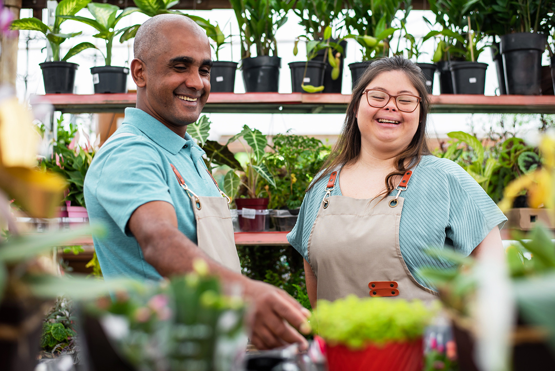 Two happy employees working in a garden nursery are surrounded by plants.
